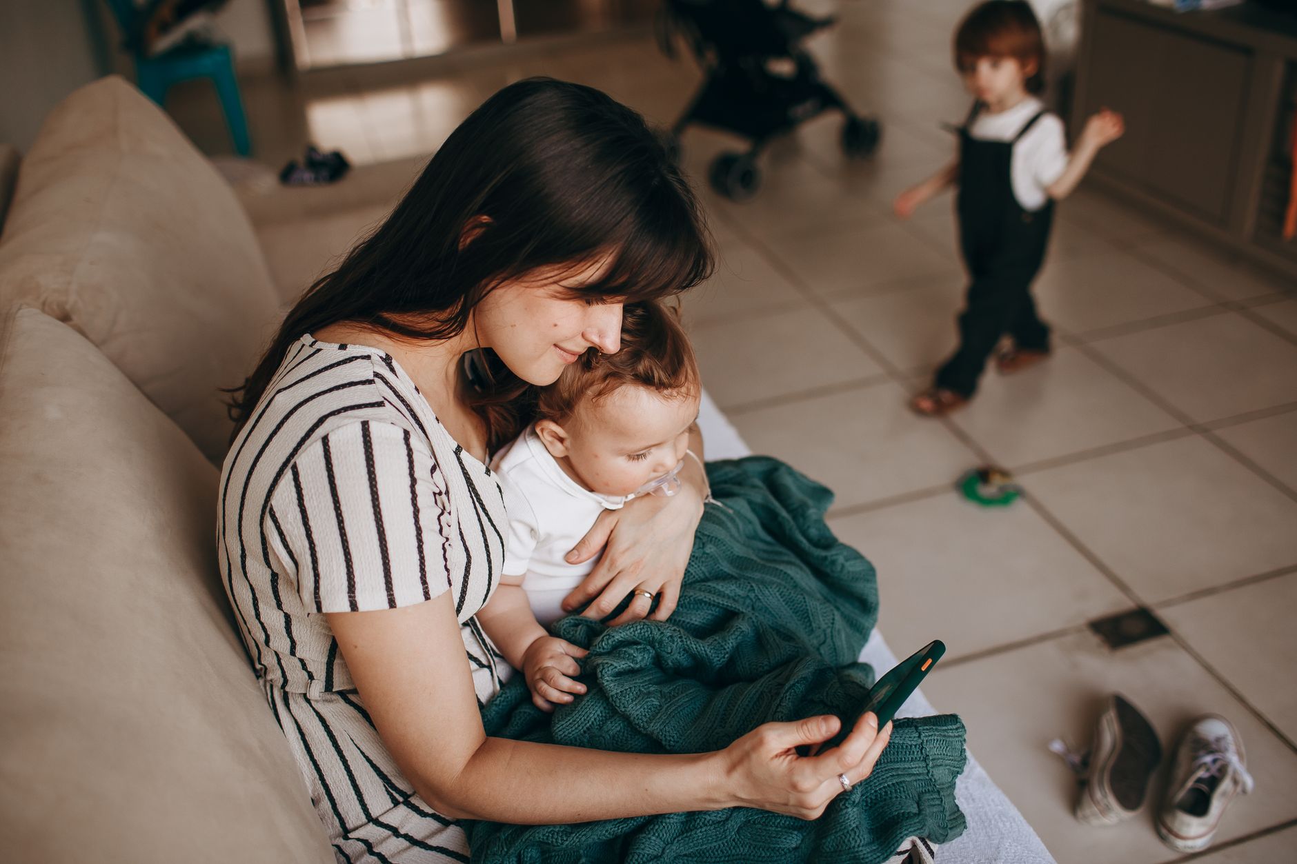 Mama z niemowlęciem na rękach przegląda telefon, starsze dziecko w tle